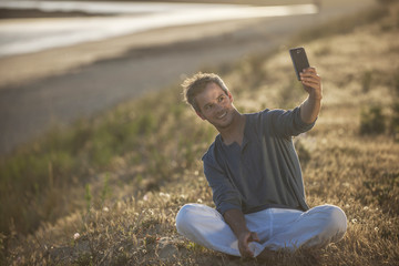 handsome man sitting on the beach at sunset and taking a selfie