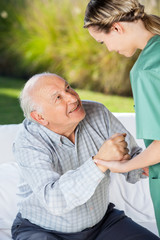 Female Caretaker Helping Senior Man To Sit On Couch