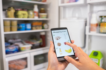 Woman makes shopping list on phone connected to the refrigerator