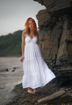 Beautiful redhead woman in white dress goes along the coast.