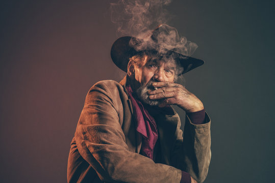 Old rough western cowboy with gray beard and brown hat smoking a