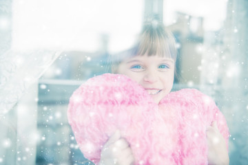 Little girl holding a heart shaped pillow and smiling behind a w