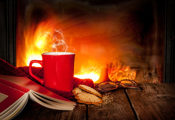 Hot tea or coffee in a red mug, book and fireplace