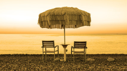 Two empty chairs stand on beach under open umbrella overlooking