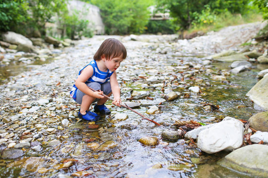 lovely toddler boy plays with stick in flashy river