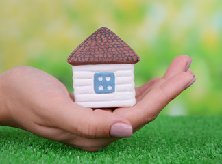 Woman hand holding small house on grass on bright background