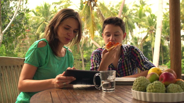 Mother with son on terrace eating, talking and using tablet comp