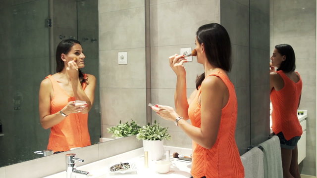 Woman applying makeup on her face with brush in front of the mir
