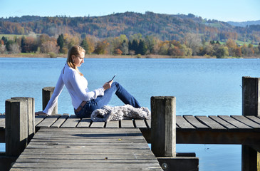 Girl reading from a tablet on the wooden jetty against a lake. S