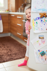 Cute little girl smiling behind refrigerator door in kitchen