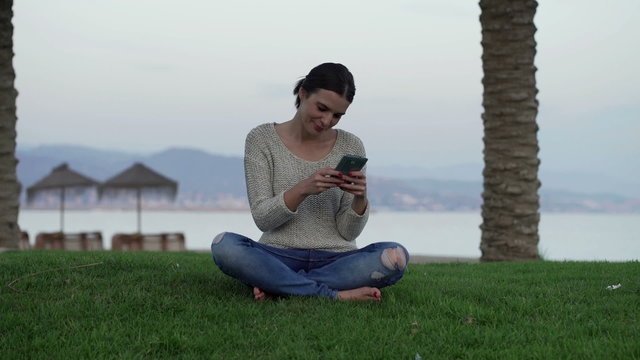 Woman texting, sending sms on smartphone sitting on grass by sea