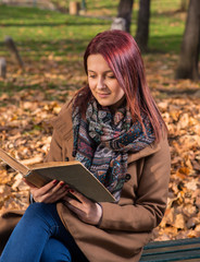 Obraz premium redhead girl sitting on bench in park and reading book