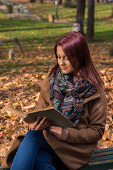 Obraz premium redhead girl sitting on bench in park and reading book