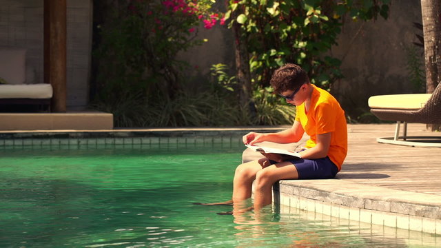 Teenager sitting with book on the edge of swimming pool