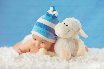 Cmile baby in hat, hugging toy on a white bedspread, on a blue b