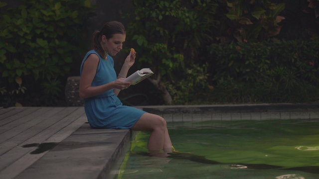 Young woman reading book on the edge of swimming pool