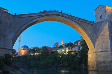 Illuminated old Ottoman bridge in Mostar at dusk with buildings of Čaršija and the mosque in the...