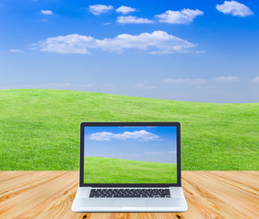 Laptop computer on wooden floor with green fields and blue sky b