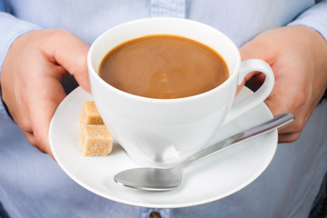 Hand holds mug of coffee with milk with spoon and brown sugar