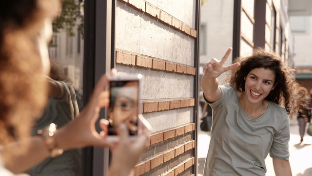 Young woman uses smartphone to take photo of friend waving in st