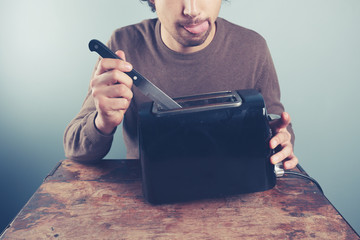 Young man sticking knife in toaster - obrazy, fototapety, plakaty