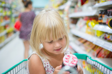 Adorable girl sit with set of good in shopping cart in supermark