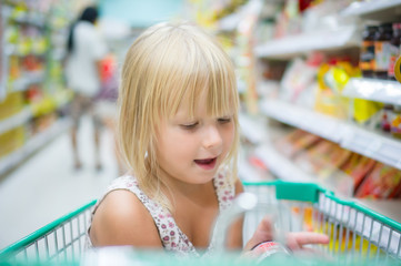 Adorable girl sit with set of good in shopping cart in supermark