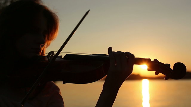 Girl violinist playing the violin at sunset on the lake.