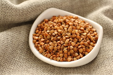 Buckwheat grains in white ceramic bowl on sackcloth background