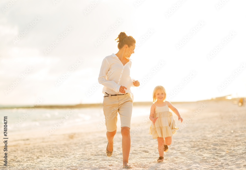 Poster happy mother and baby girl running on beach at the evening