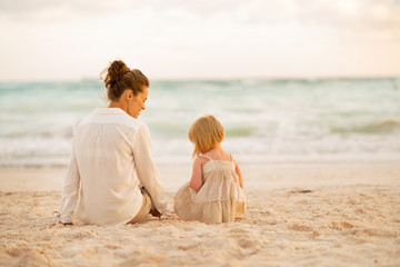 Mother and baby girl sitting on beach at the evening. rear view