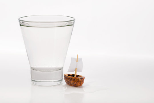 Closeup of a walnut shell boat with a sail, close to a transparent glass full of water or alcohol