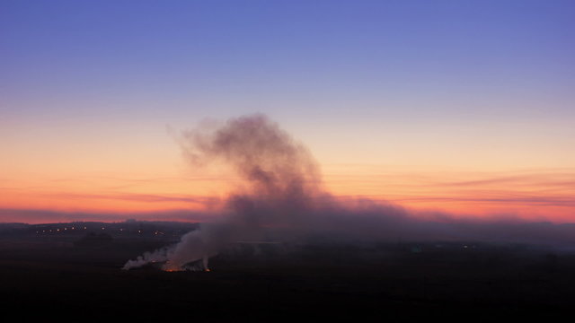 The smoke in the field near the highway and industrial objects