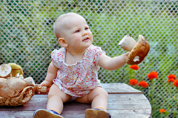 Baby girl sits on the wooden table and holding boletus