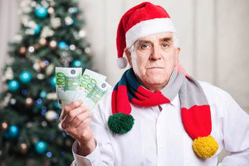 Senior man in Santa hat holding money on Christmas background