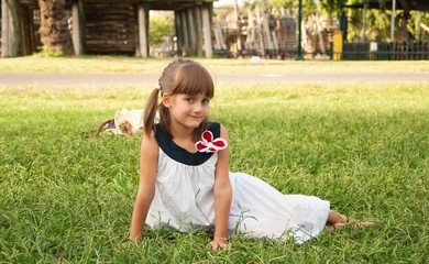 Fototapeta na wymiar Cute little girl in white dress sitting on lawn in park