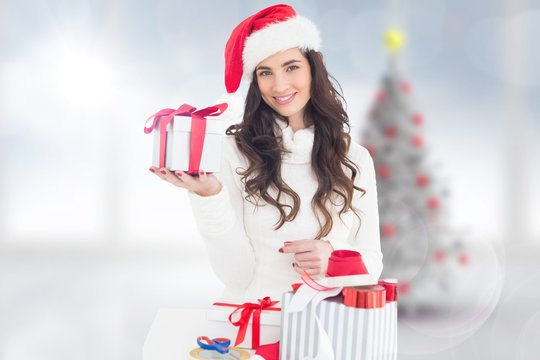 Composite image of cheerful brunette in santa hat holding gift
