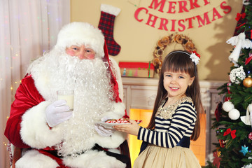 Obraz premium Little cute girl giving cookies and milk to Santa Claus near
