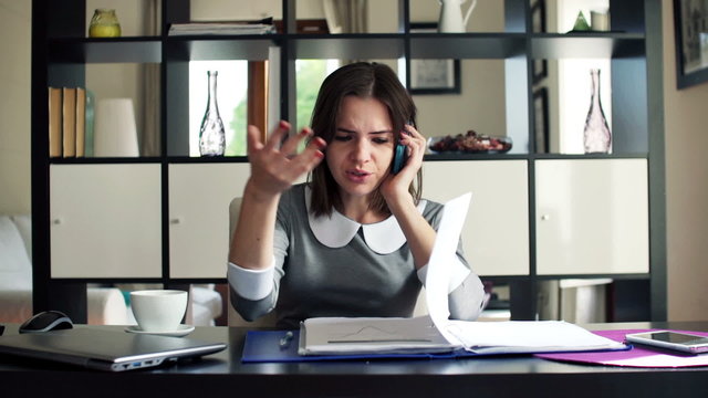 Angry businesswoman with documents talking on cellphone at home