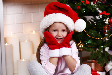 Obraz na płótnie Canvas Little girl in Santa hat and mittens sitting near fir tree