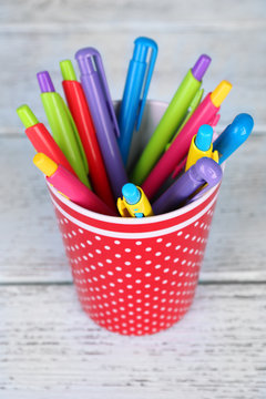Plastic cup with different pens on color wooden background