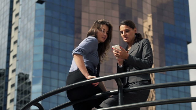 Two pretty businesswomen talking and using smartphone by glass