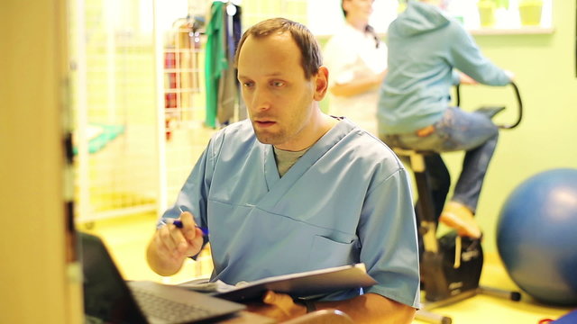 Male doctor writing notes with laptop in rehabilitation room