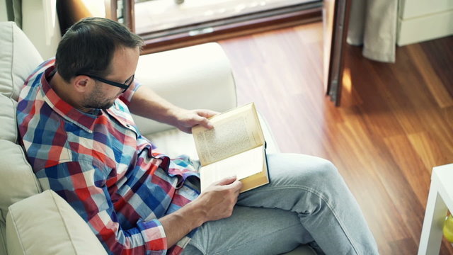 Young man reading book sitting on sofa at home, top view