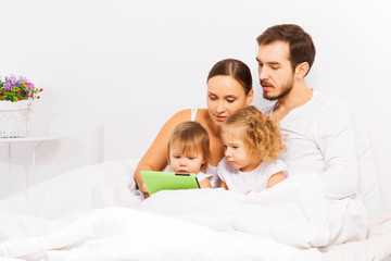 Fototapeta premium Parents and two cute kids play with tablet on bed