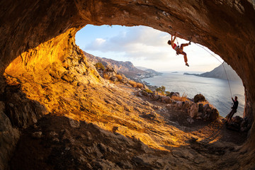 Young woman lead climbing in cave, male climber belaying - obrazy, fototapety, plakaty