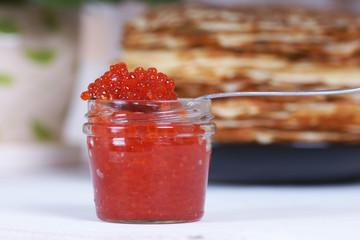 Банка красной икры на фоне большой стопки блинов на столе