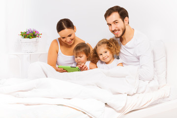 Fototapeta premium Parents and two kids play with tablet on white bed