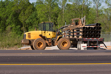 Obraz premium Getting ready to install sewer lines, a tractor trailer pulls up alongside the road so it can be unloaded by a large tractor with forks instead of a shovel. Copy space in trees or on pavement.