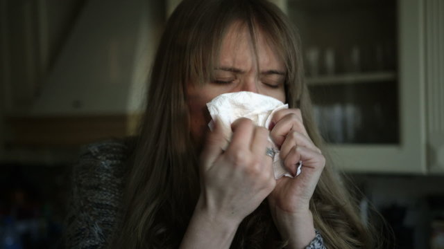Woman with a bad cold and blowing her nose in the kitchen.